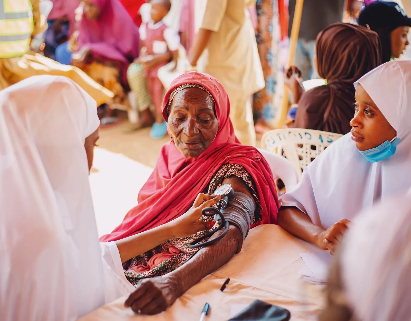 Nurse in a white hijab taking an elderly woman's blood pressure in warm light